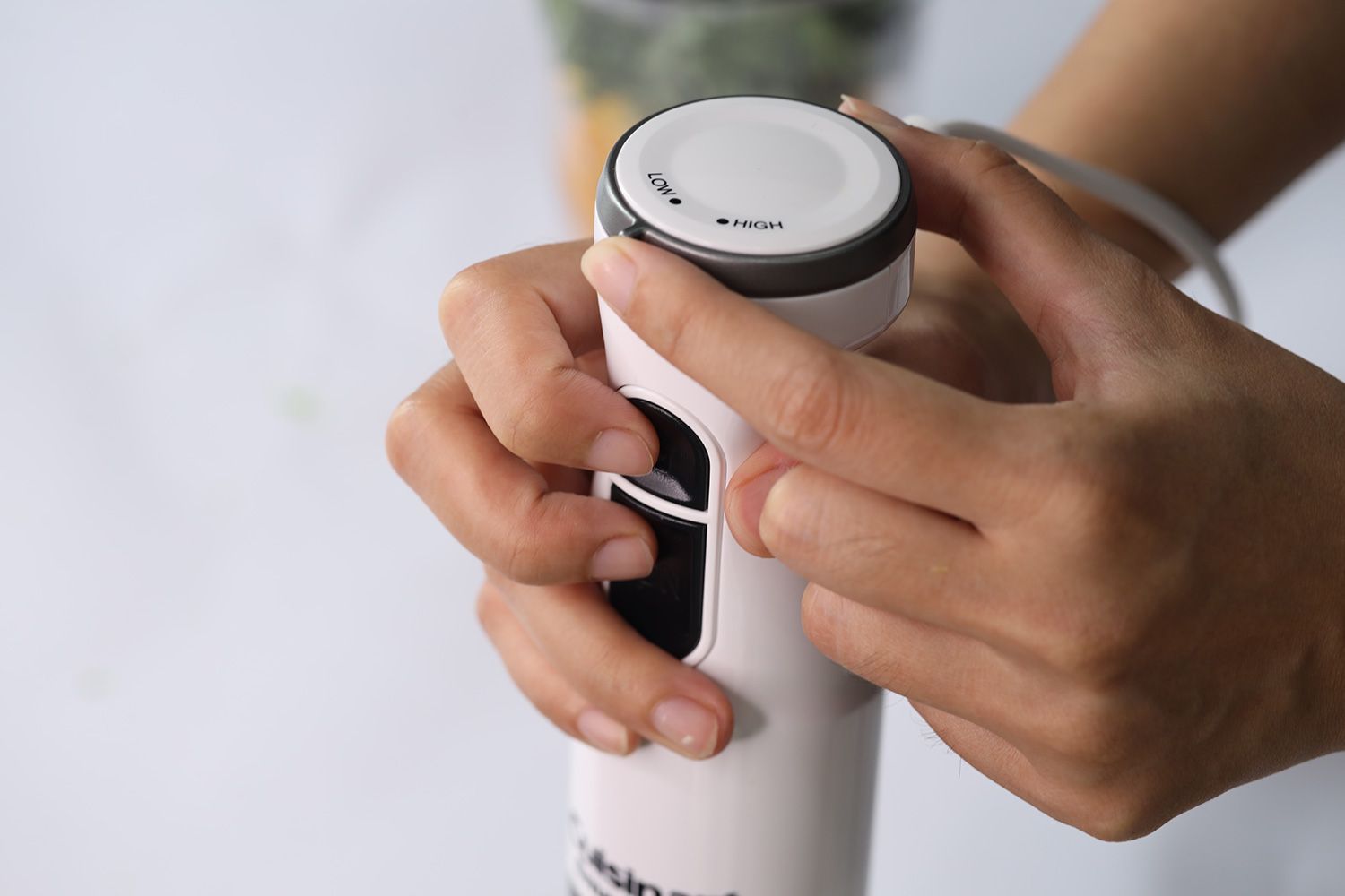 Someone uses two hands to control the Cuisinart Smart Stick immersion blender: one for holding the handle and pressing the controlling buttons at the same time, the other for rotating the speed dial. 