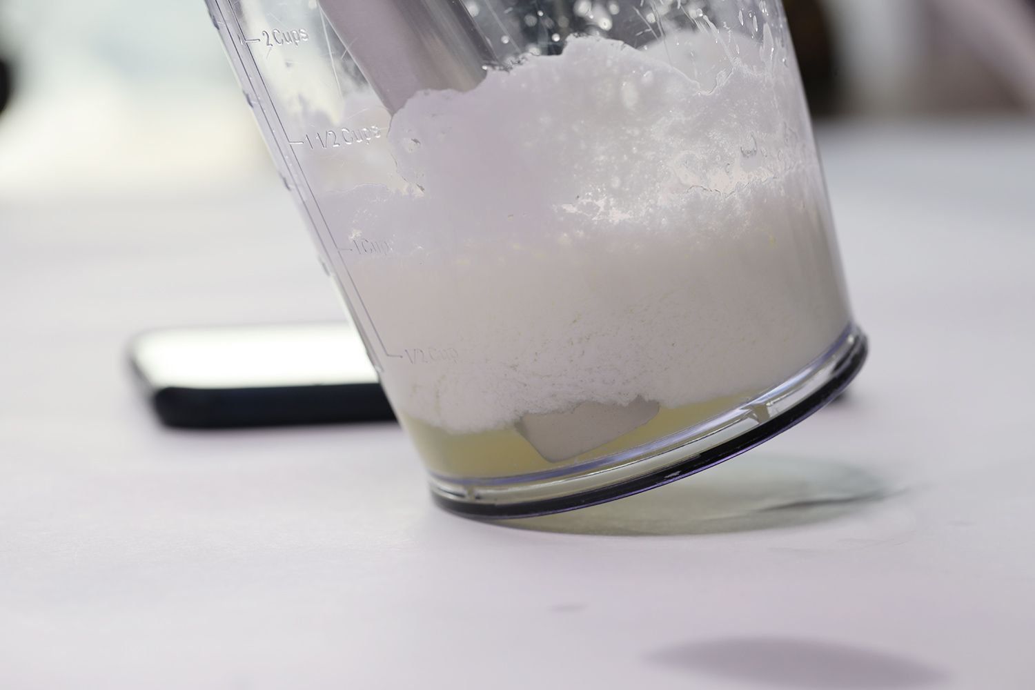 The blending wand of the Cuisinart CSB-175 Smart Stick  is immersed in a plastic beaker on a white table containing its failed beaten egg-white with a smartphone next to it.