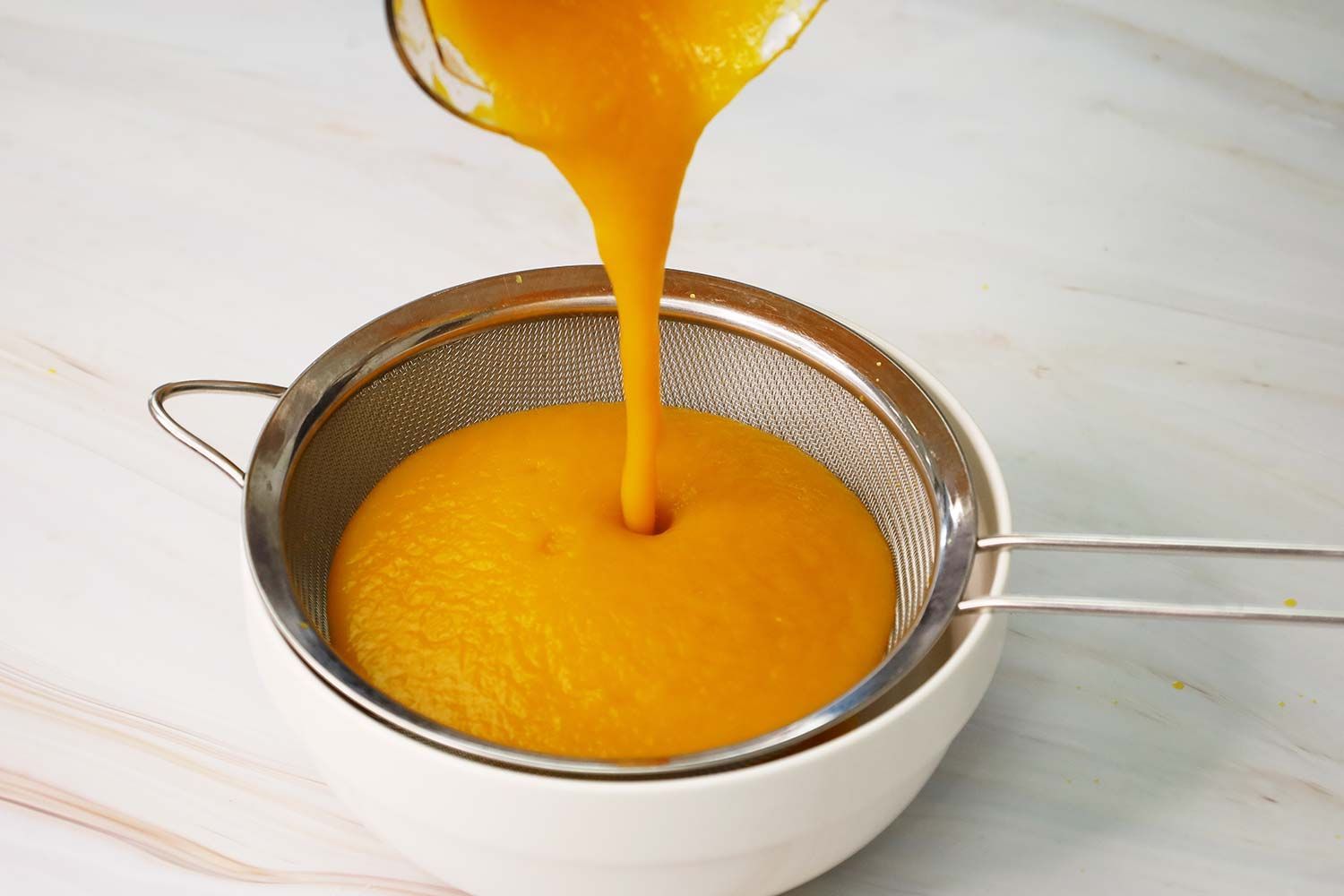 Someone pouring the pureéd soup from the plastic beaker to the stainless steel mesh strainer lying above a white bowl to check its smoothness.