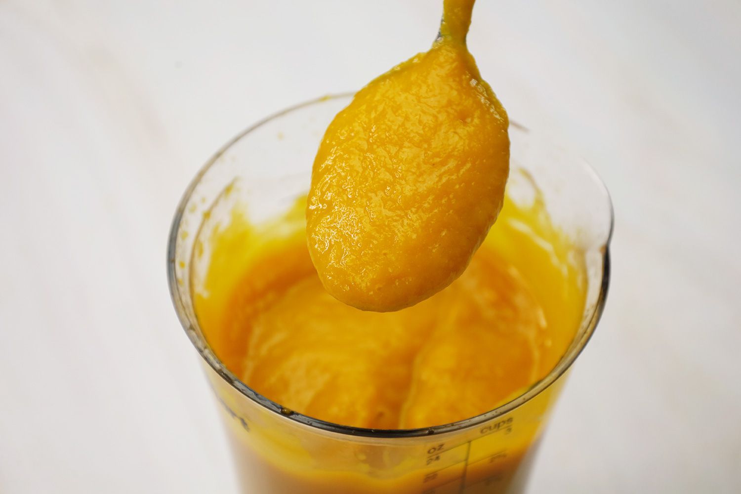 Scooping a spoon of pureéd soup prepared by the KitchenAid battery-powered immersion blender from the full batch in the plastic beaker. 