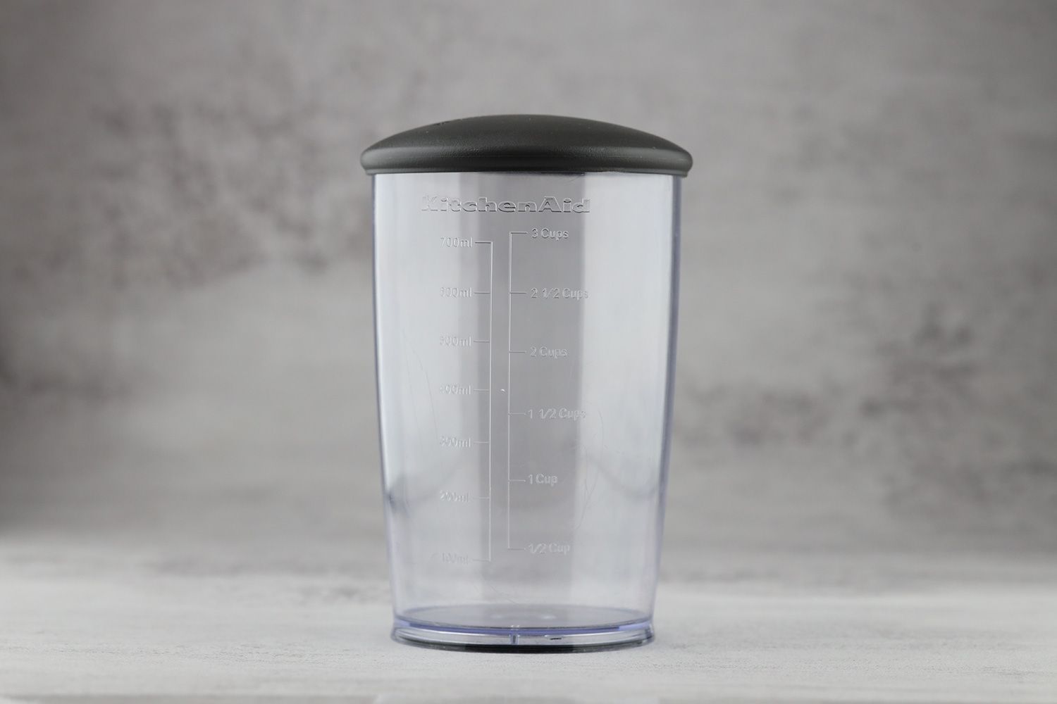 An included 700 ml plastic beaker of the KitchenAid cordless immersion blender standing on a gray table. 