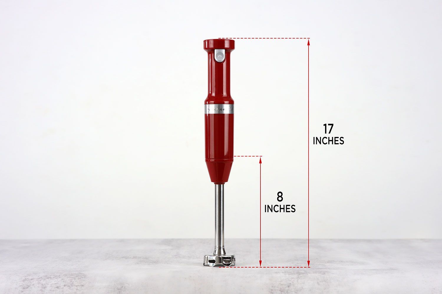 The KitchenAid cordless hand blender standing on top of its blending shaft on a gray table, with the length of the blending shaft being noted to the side as 8 inches, and the total length of the unit as 17 inches.