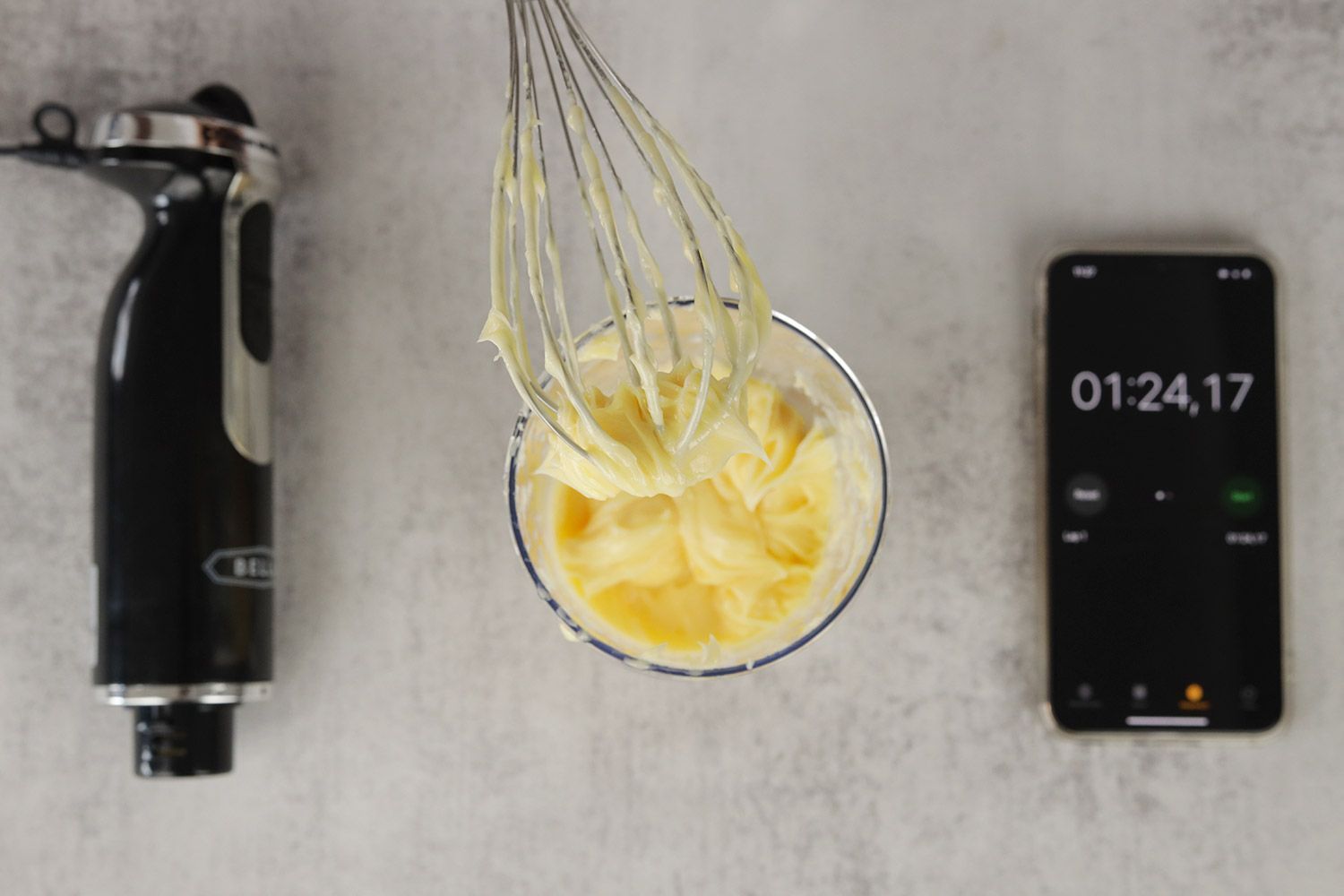 A little bit of mayonnaise sticking in the whisk attachment of the BELLA hand blender when it is removed from the full batch emulsified in a plastic beaker standing between the BELLA motor body and a smartphone displaying the total emulsion time (1 minute and 24 seconds). 