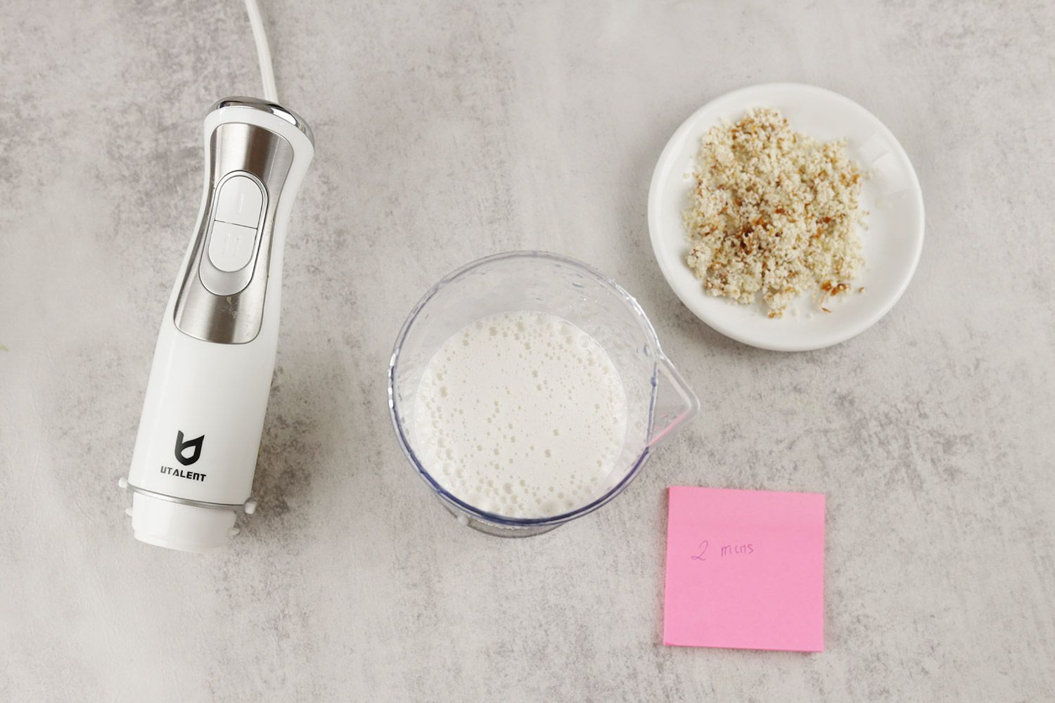 The UTALENT’s motor body, a plastic beaker containing almond milk, a white plate of almond pulp, and a small red note displaying the total grinding time (2 minutes) being side by side on a gray table. 