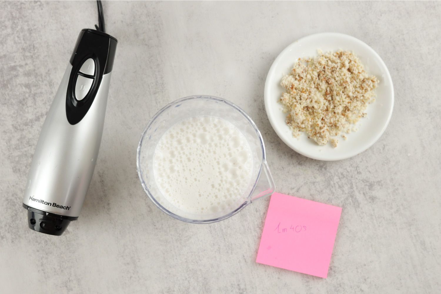 The Hamilton Beach motor body, a plastic beaker containing almond milk, a white plate of almond pulp, and a small red note displaying the total grinding time (1 minute 40 seconds) being side by side. 