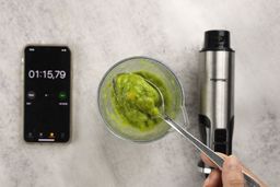 A plastic beaker containing a batch of green smoothie whose parts are scooped with a stainless steel spoon is between the Mueller’s motor body and a smartphone displaying the total blending time (1 minute and 15 seconds).