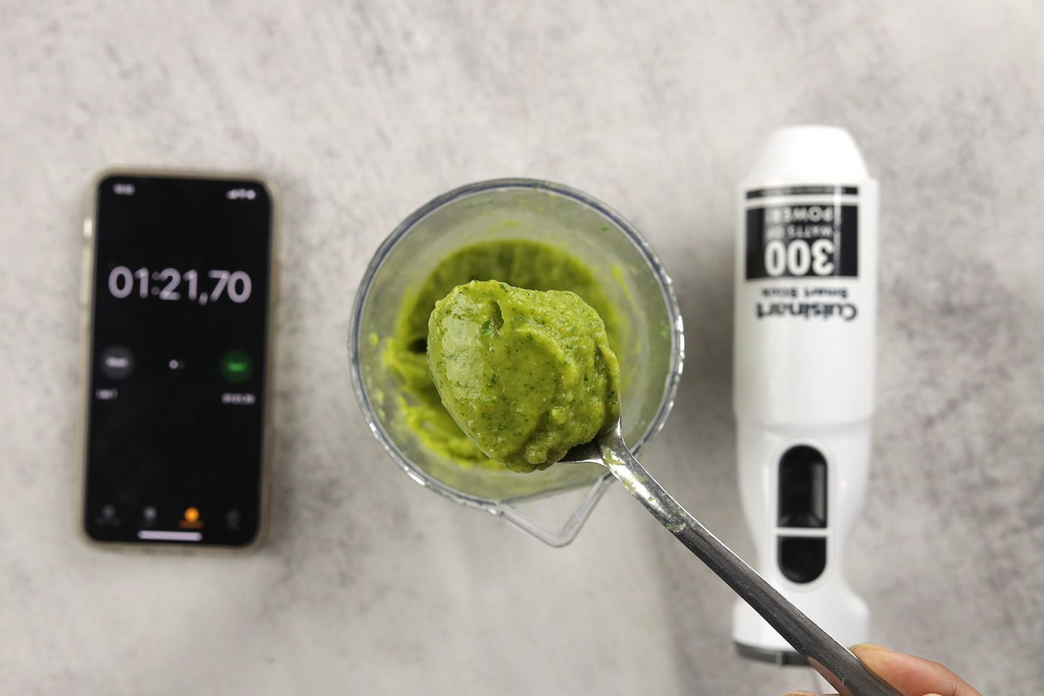 A plastic beaker containing a batch of green smoothie whose parts are scooped with a stainless steel spoon is between the Cuisinart’s motor body and a smartphone displaying the total blending time (1 minute and 21 seconds).