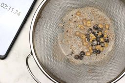 A batch of protein shake packed with dried blueberries, oatmeal, and almonds prepared by the Magic Bullet Personal Blender is checked for smoothness by being drained through a stainless steel mesh strainer, with a smartphone displaying the total blending time (2 minutes and 11 seconds) next to it.