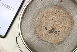 A batch of protein shake prepared by the Oster Myblend Single-Serve Blender is checked for smoothness by being drained through a stainless steel mesh strainer, with a smartphone displaying the total blending time (2 minutes and 50 seconds) next to it. 