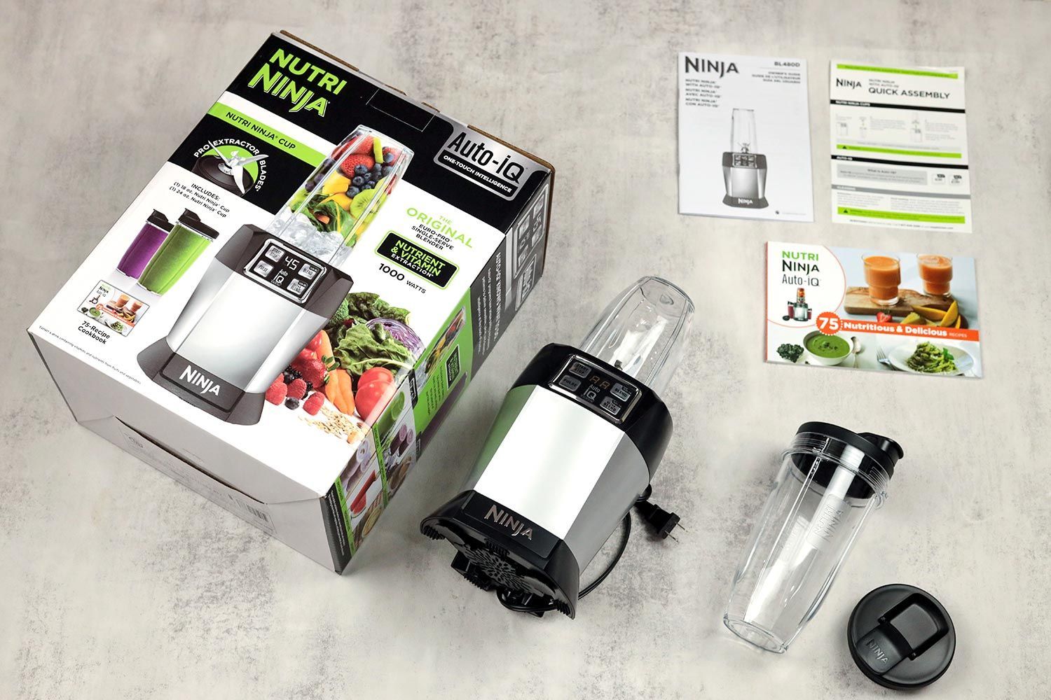 The Ninja Nutri Auto-IQ 1000-Watt lying on a gray table with its accessories, including two to-go lids, an additional blending cup, a recipe book & user guide, and a paper carton box, by its sides.