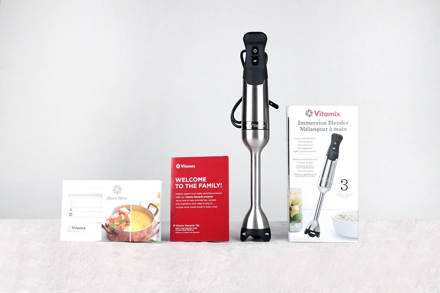 Unboxing the Vitamix immersion blender, from left to right: an owner's manual, a recipes booklet, a red receipt, the Vitamix 5-speed with a stainless steel motor body and an immersion blending wand, and a paper carton box.