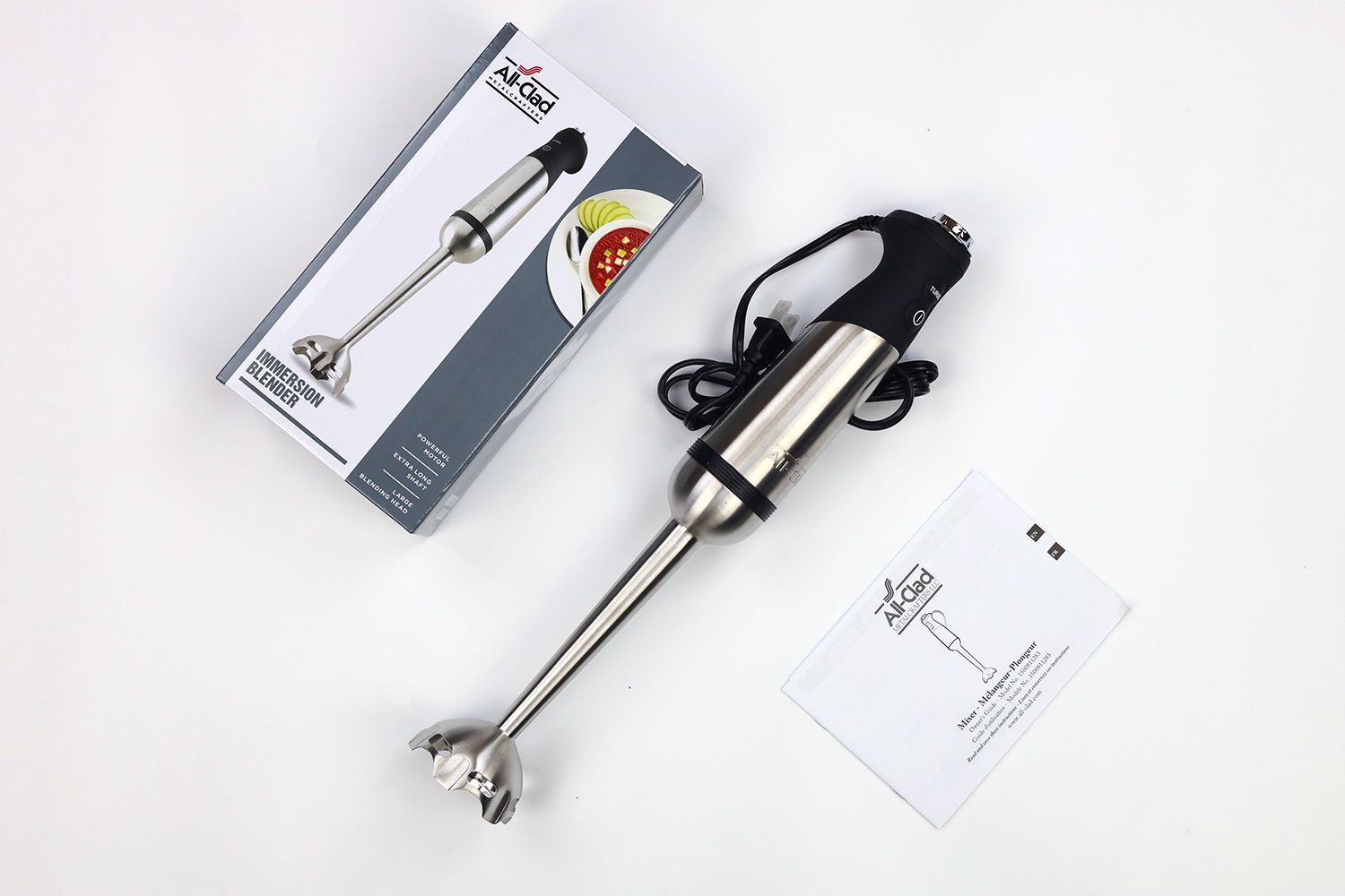 All-Clad immersion blender review: a paper carton box, the All-Clad blending wand with its motor body attached, and a user’s manual lying side by side on a white table.