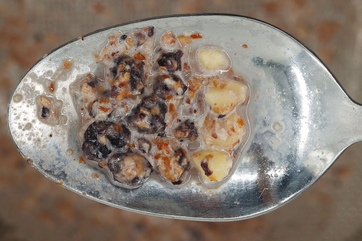 Using a spoon to remove the unblended blueberries chunks, including almonds and dried blueberries, from a mesh strainer. 