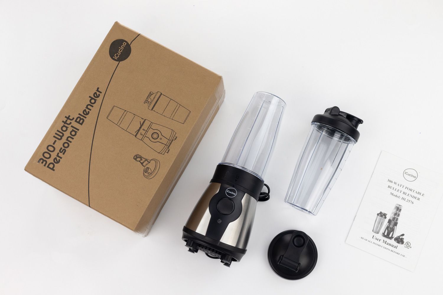 The iCucina Personal Blender with a motor base and blending cup attached lying on a table with a to-go lid, a blending cup with lid, a paper carton box, and a user guide by its sides.