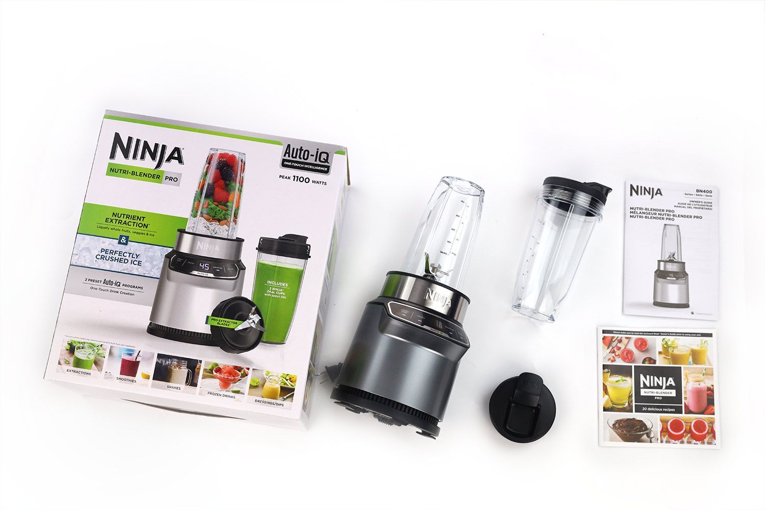 The Ninja BN401 Nutri Pro personal blender lying on a white table with the accessories by its side, including a blending cup with lid, a spout lid, a paper carton box, a recipe book, and owner’s manuals.