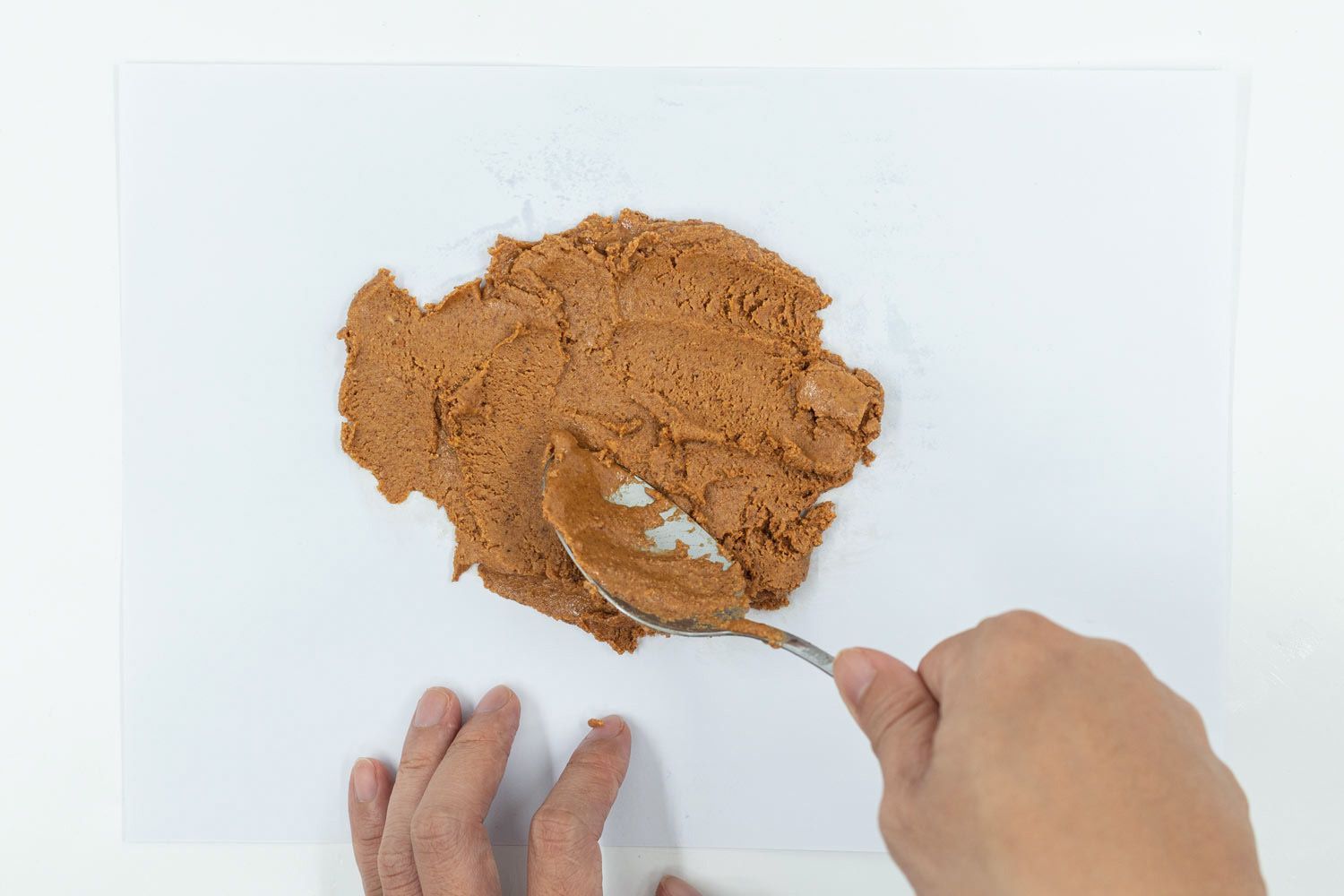 Someone is extracting a spoonful of almond butter and apply it evenly on a white sheet of paper to examine its texture.