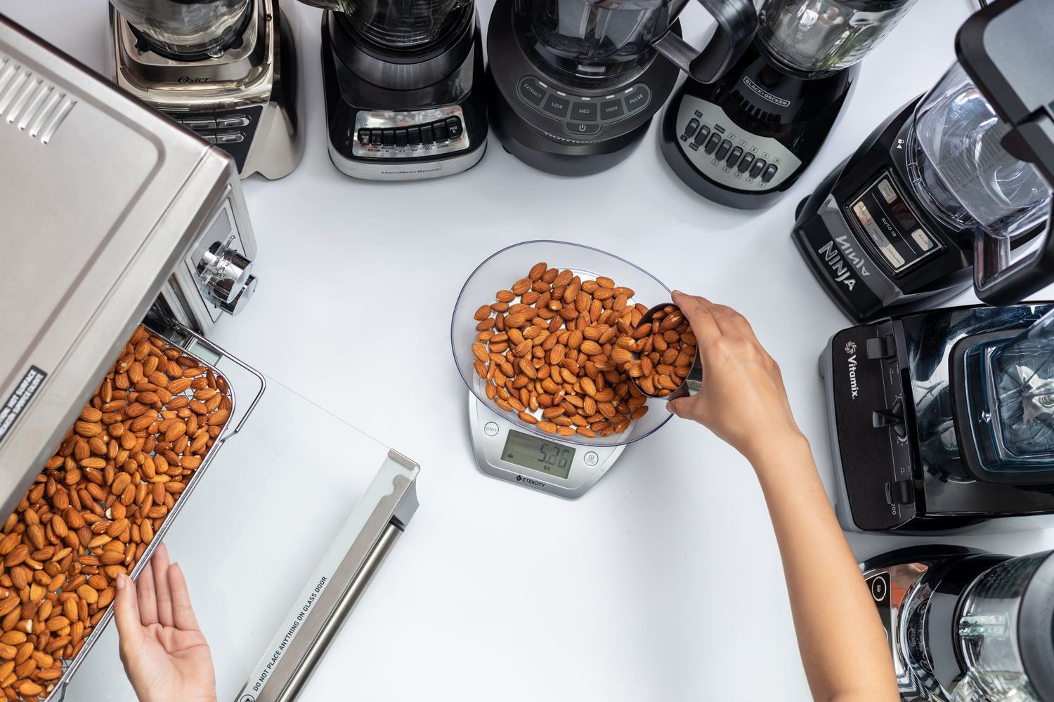 Someone is preparing for the almond butter test with the tray of almonds, a metal cup, and a scale nearby. The Vitamix blender's motor, tamper, and lid are also present, along with a smartphone for timing and recording the blending process.