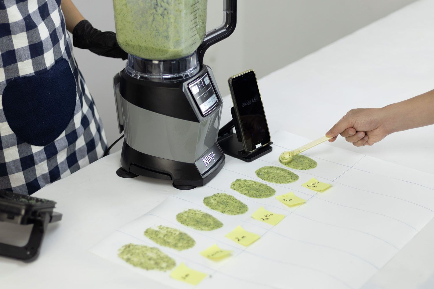 Tina is holding the Ninja AMZ493BRN Compact Kitchen System blender, with a smartphone sitting next to it on a countertop. Lap Vo is using a spoon to spread a small amount of green smoothie onto a white piece of paper.