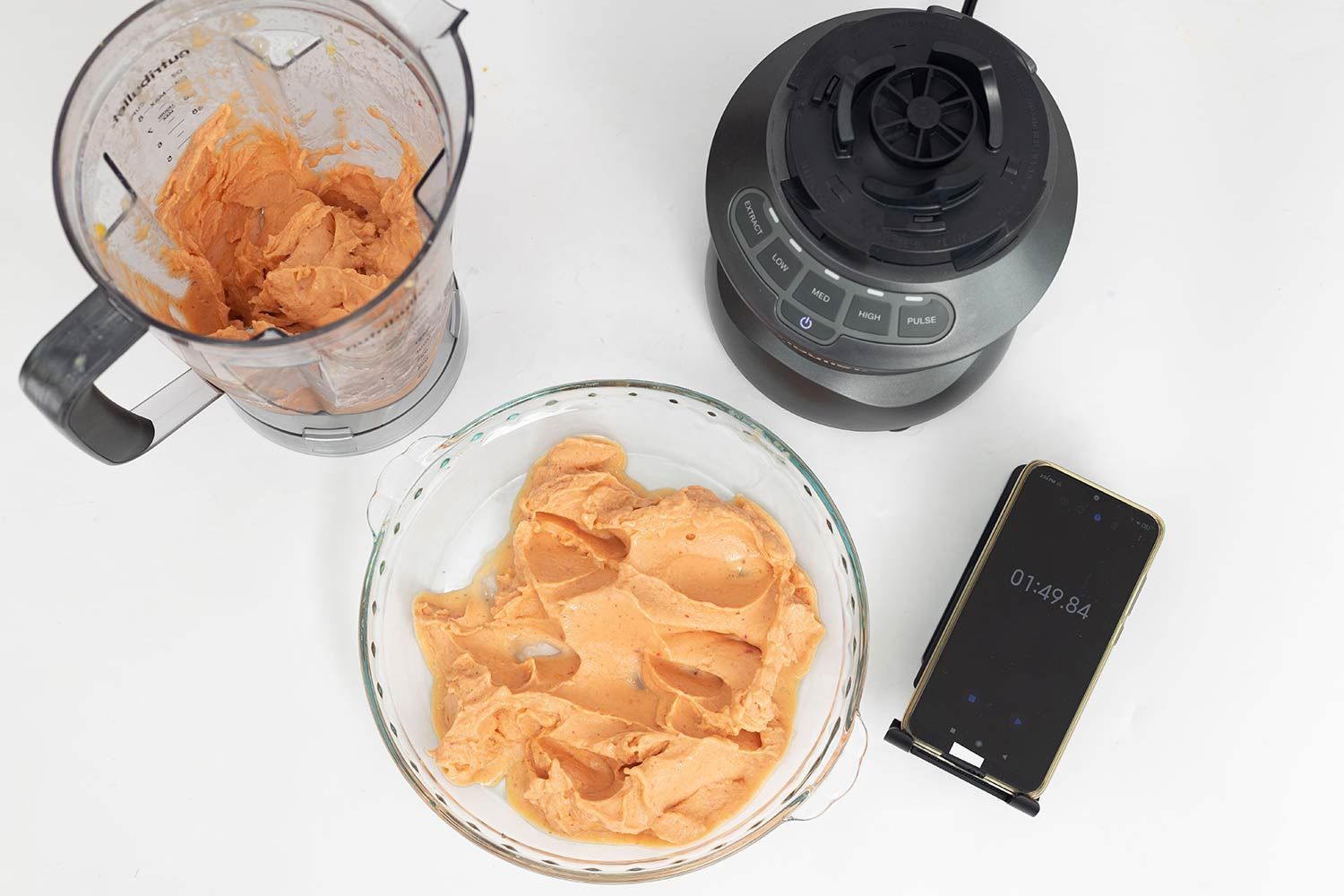 The NutriBullet motor base stands beside the container, which holds a portion of the smoothie it produced. Next to it, a glass bowl contains the remaining smoothie, while a smartphone displays a blending time of 1 minute and 49 seconds.