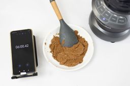 The NutriBullet is beside a white plate containing almond butter with a spatula nestled inside, and a smartphone revealing a blending time of 6 minutes.