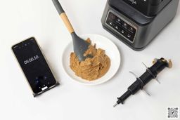 The Ninja BN701 Professional Plus is beside a white plate containing almond butter with a spatula, blend assembly, and a smartphone revealing a blending time of 5 minutes.