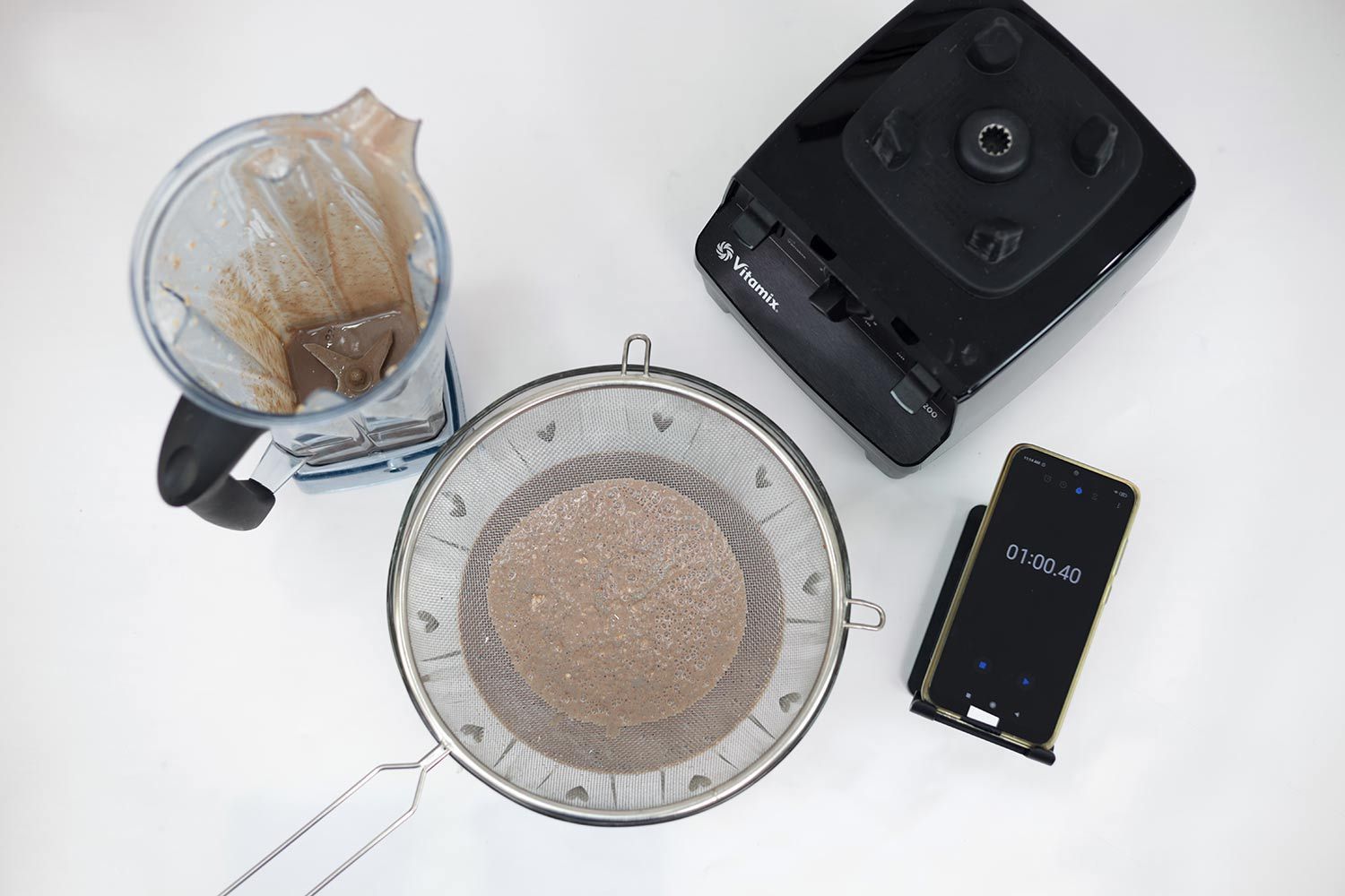 A Vitamix blender setup with residue in its jar, a sieve with strained protein shake beside it, and a smartphone displaying a timer of "01:00.40" on a white surface. 