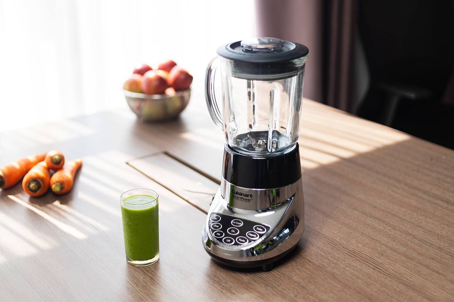 The Cuisinart blender on a kitchen counter with a green smoothie next to it, with a background of a bowl of apples and carrots.