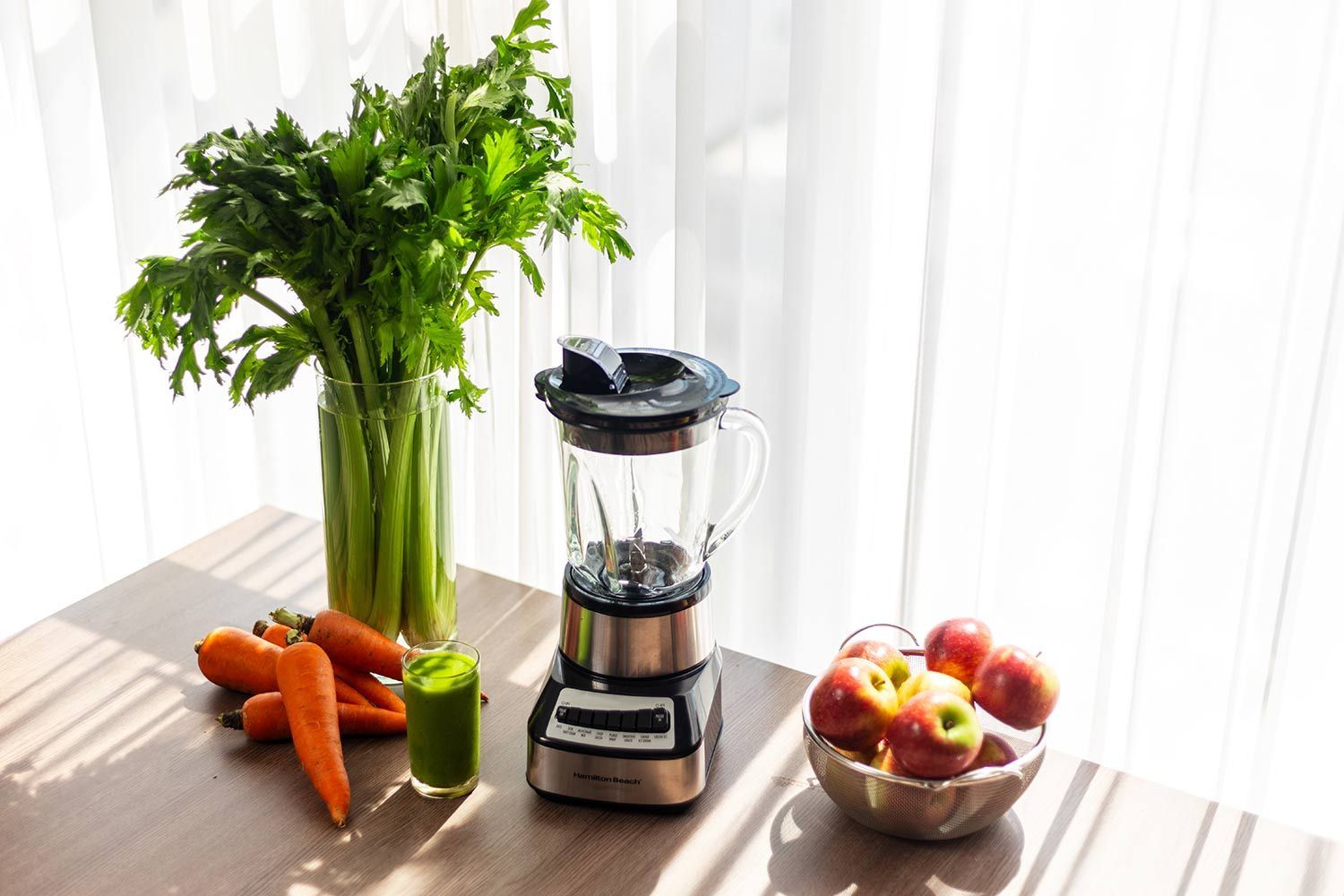 The Hamilton Beach blender on a kitchen counter, near a glass of green smoothie, a bunch of celery, and a bowl of apples.