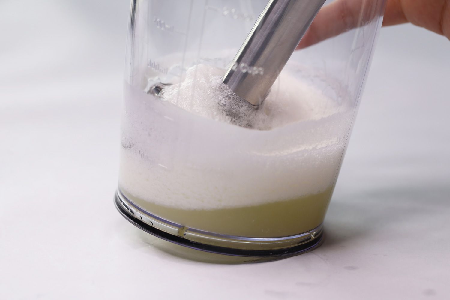 The blending wand of the KitchenAid KHBBV53 is immersed in a plastic beaker on a white table containing its failed beaten egg-white.