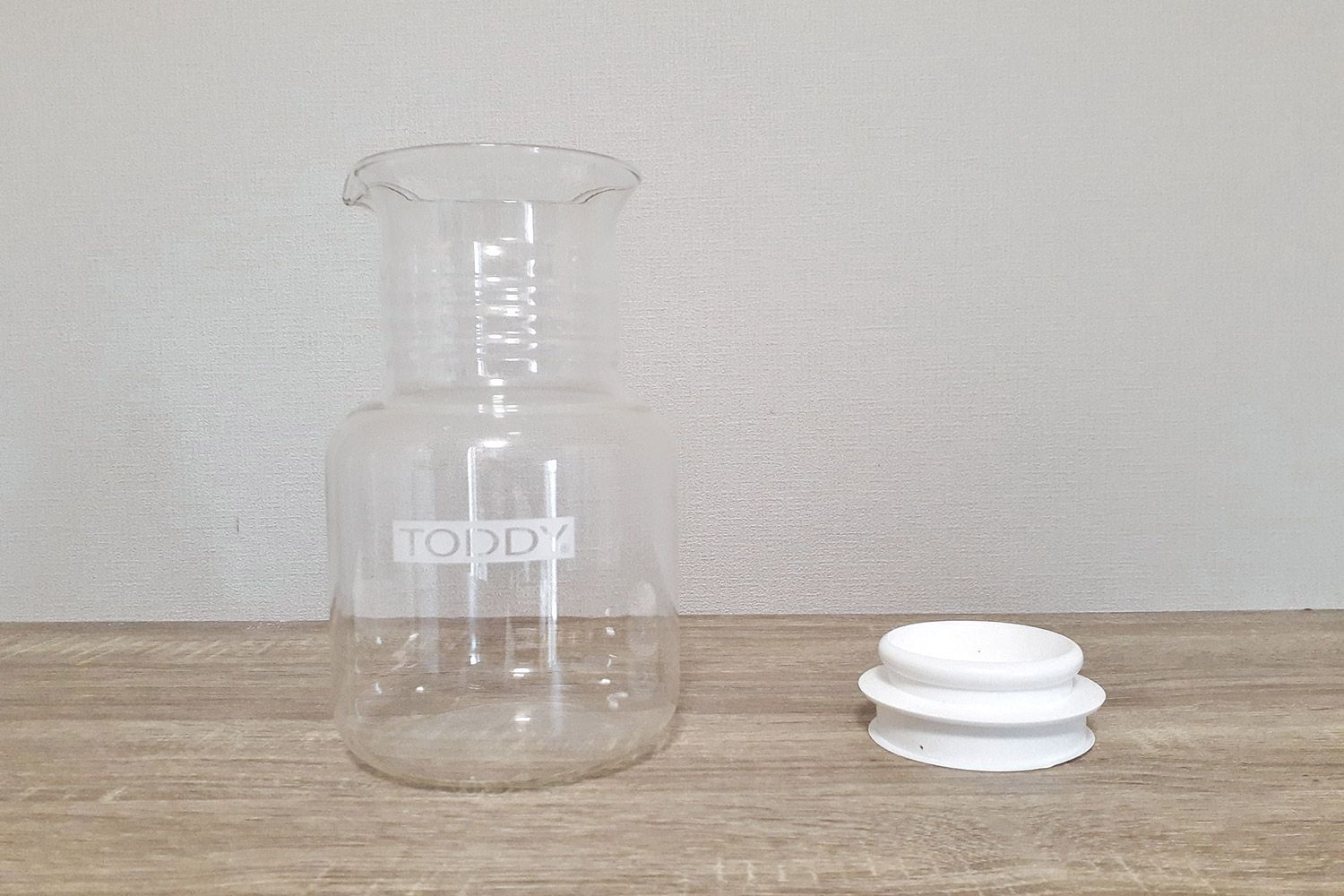 The glass carafe of the Toddy Cold Brew System on a wooden counter with the carafe lid to one side.