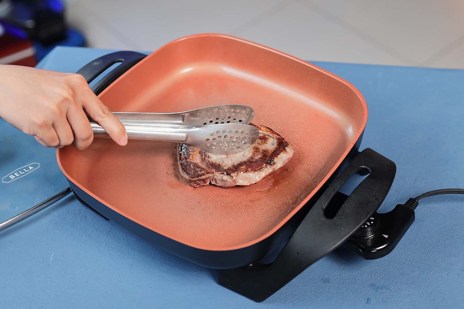 A person holding a steak with a tong to sear the edge of a steak in the BELLA Electric Skillet 14607.