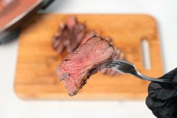 A hand in black glove using a fork to pick up a slice of steak. In the background is the Bella Non-Stick Electric Skillet 14607.