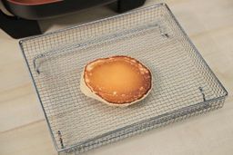 The top side of a golden brown pancake that’s dark around the edges inside an air fryer basket. In the corner is the Bella Non-Stick Electric Skillet 14607.