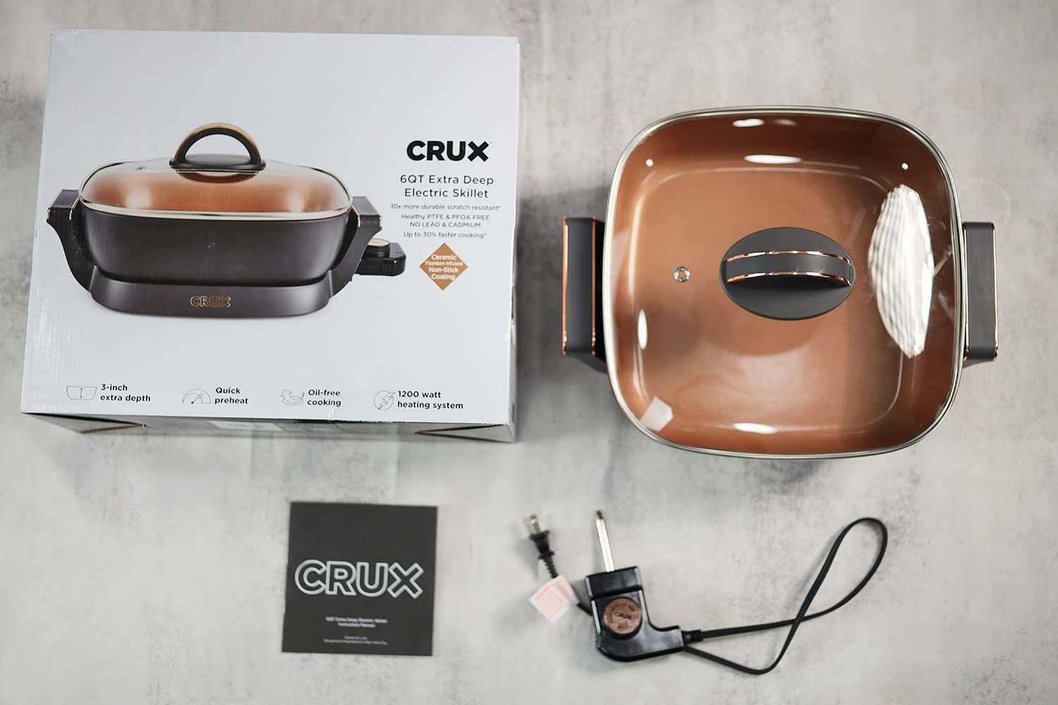 On the upper right is the Crux Non-Stick Electric Skillet 14620. On the left is its cardboard box. Below the skillet, on the left is a thermostat and on the right is an instruction manual.