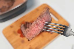 A hand in black glove using a fork to pick up a slice of steak. In the background is the Crux Non-Stick Electric Skillet 14620.