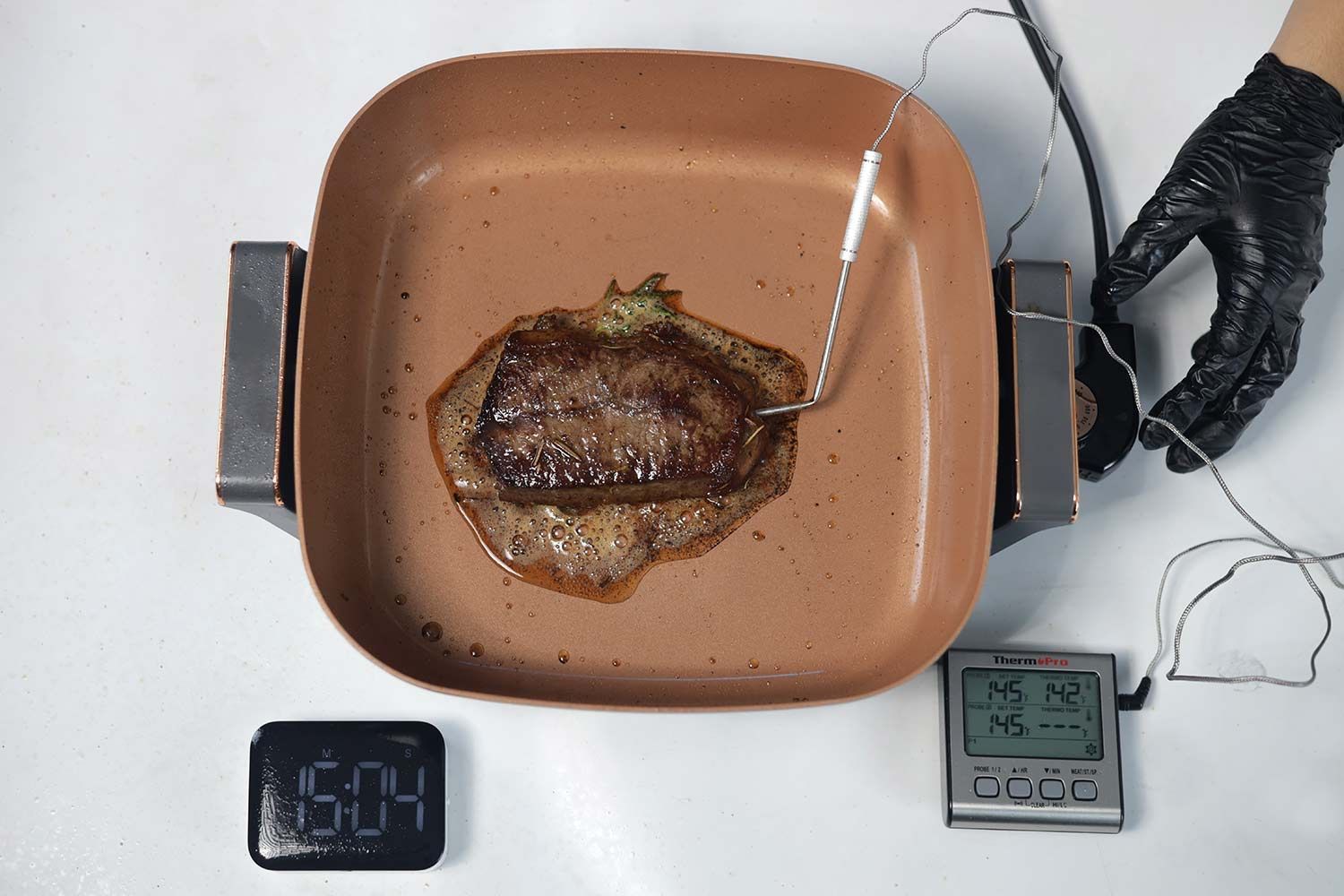 A piece of deep golden brown steak inside the Crux Non-Stick Electric Skillet 14620. In front of the skillet is a digital timer and a meat thermometer with its probe inserted into the steak.