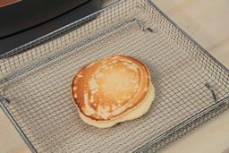 The top side of a golden brown pancake that’s dark around the edges inside an air fryer basket. In the corner is the Crux Non-Stick Electric Skillet 14620.