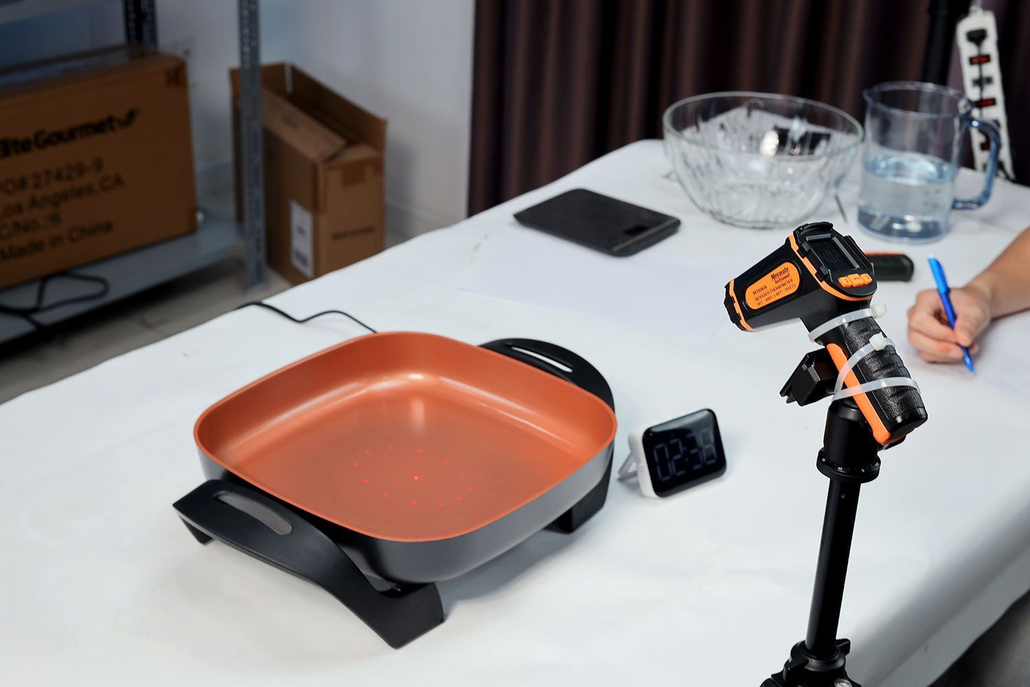 On a white table is an infrared thermometer fixed on a stand, the Bella Non-Stick Electric Skillet 14607, and a digital thermometer.