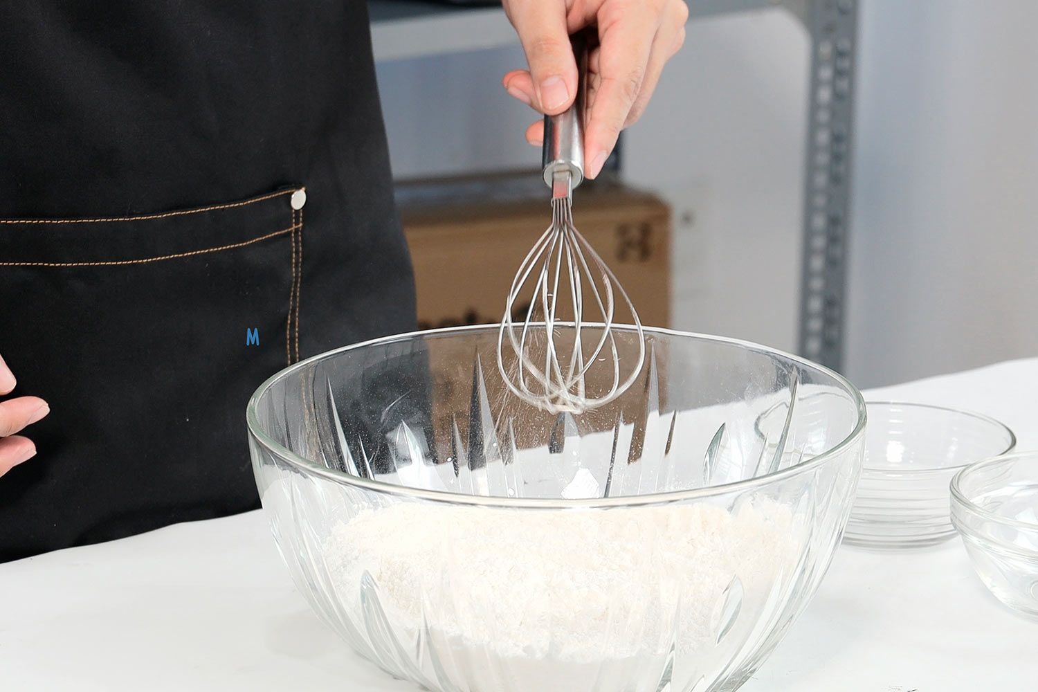 A person in an apron holding a whisk to incorporate dry ingredients.