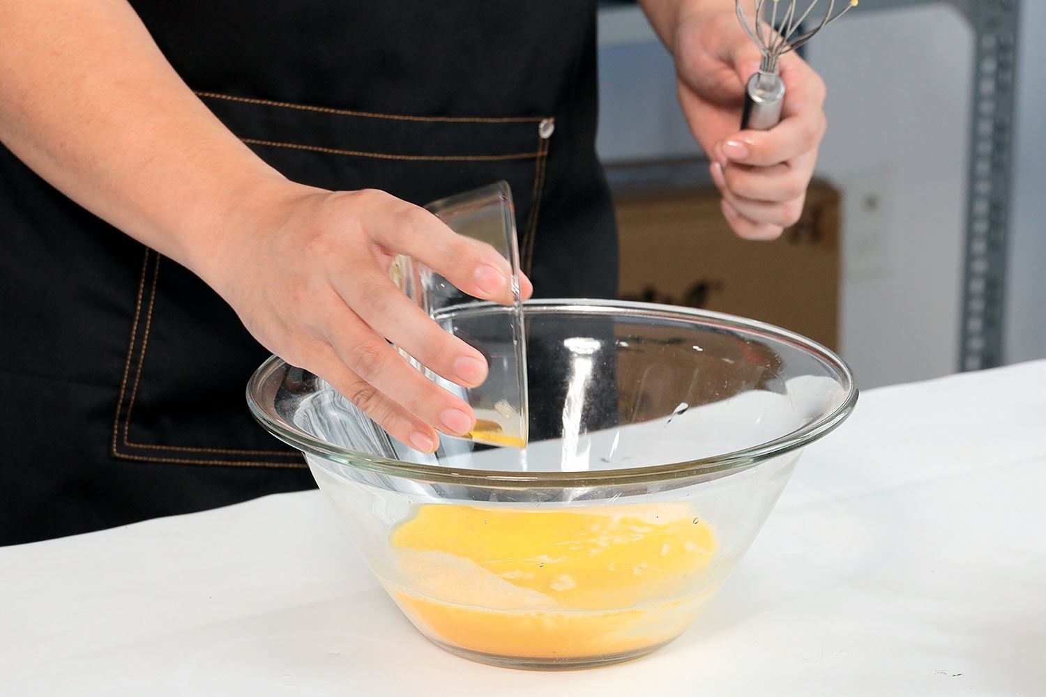 A person in an apron holding a whisk pouring butter into a bowl of wet ingredients.