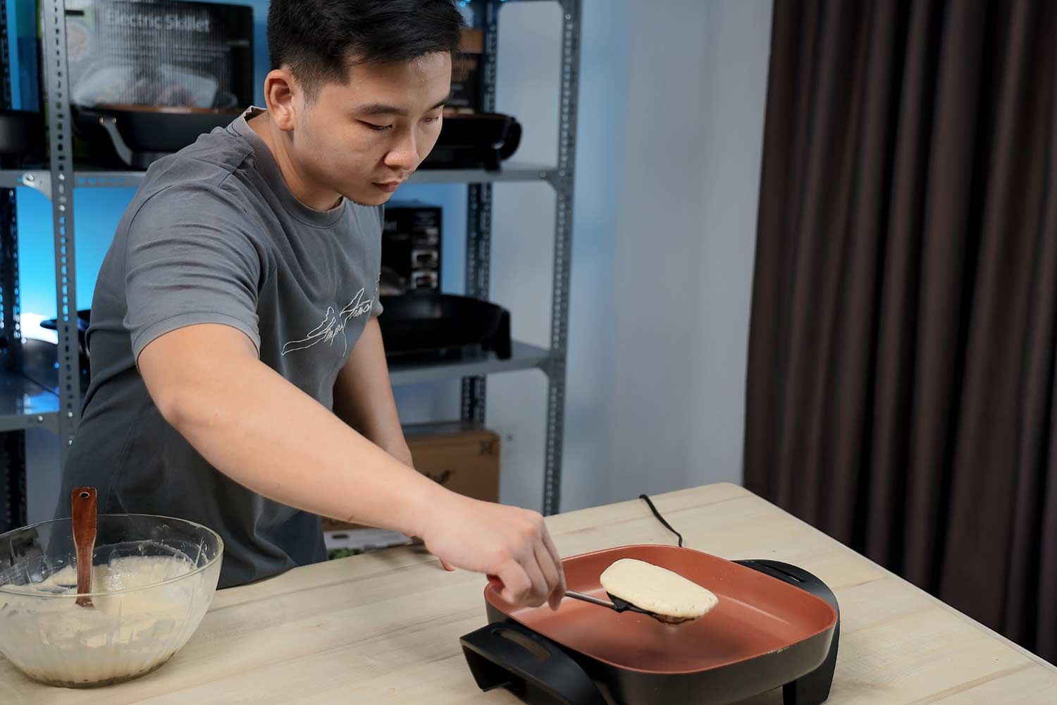 A person flipping a pancake in the Bella Non-Stick Electric Skillet 14607. Next to it is a bowl of batter.