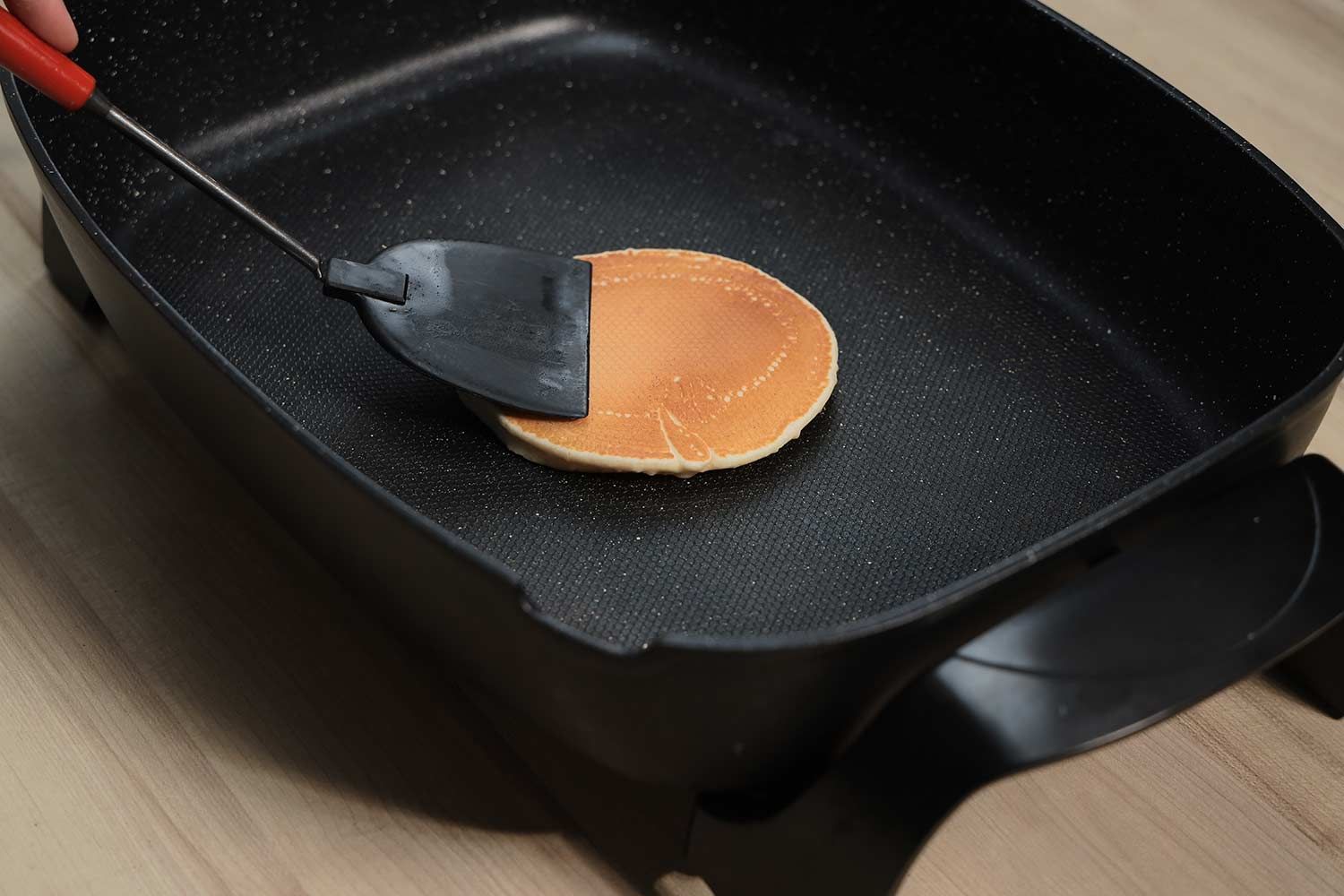 A golden brown pancake inside the Elite Gourmet Non-Stick Electric Skillet EG-6203#. A spatula is pressing down on it.