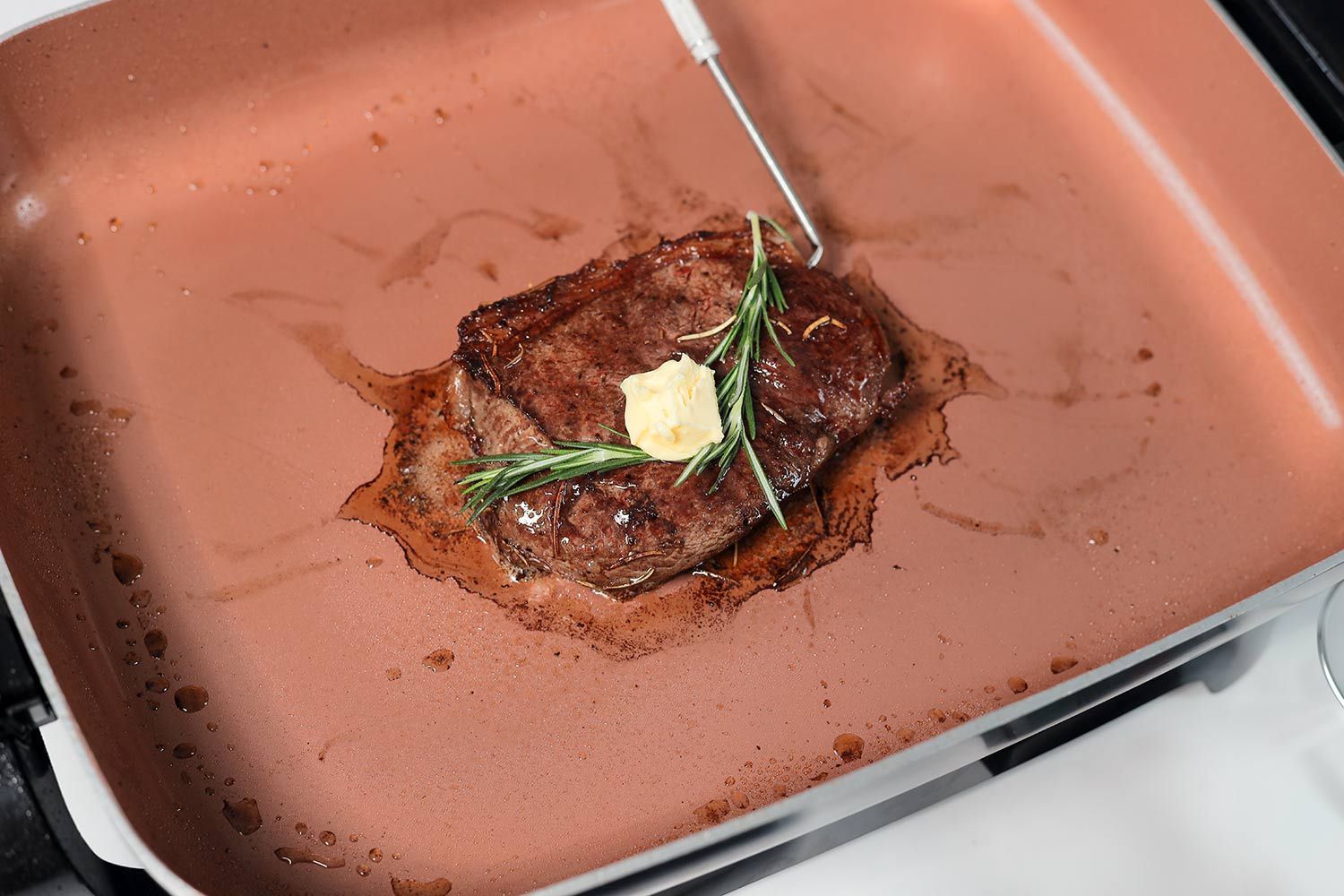 A seared steak with a probe inside, topped with a knob of butter and a twig of rosemary, inside the Hamilton Beach Ceramic Non-Stick Electric Skillet 38529K.