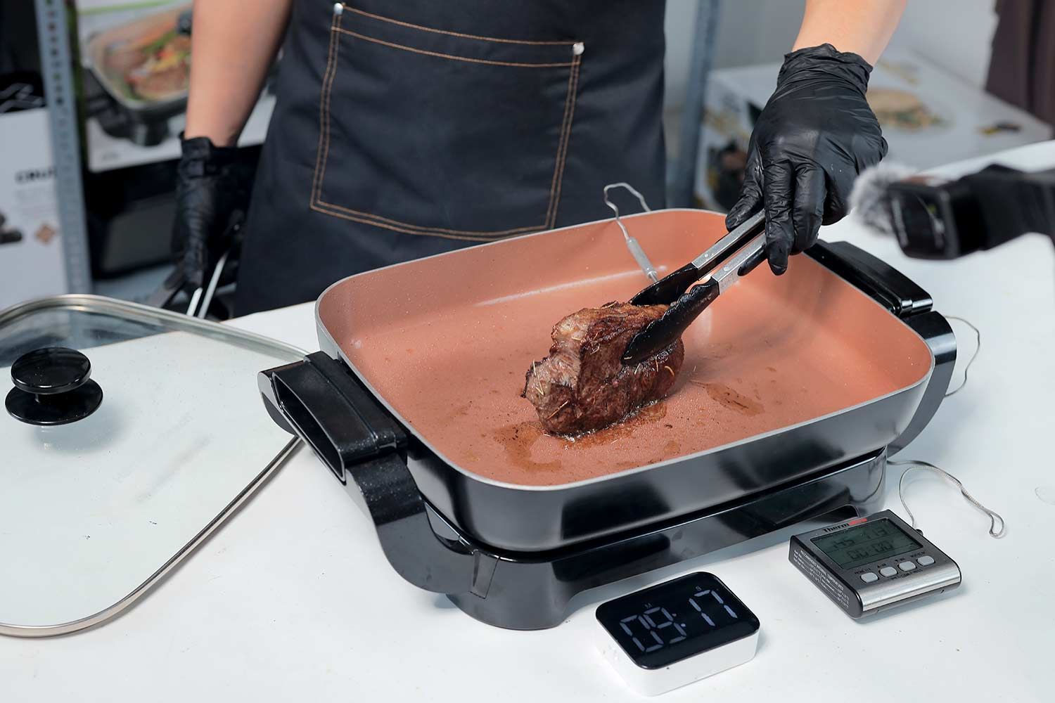 A person holding a steak with a tong to sear the edge of a steak in the Hamilton Beach Ceramic Non-Stick Electric Skillet 38529K. A digital timer, and a meat thermometer.