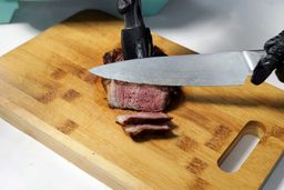 On the cutting board, a tong is holding the steak while a knife is slicing the steak.