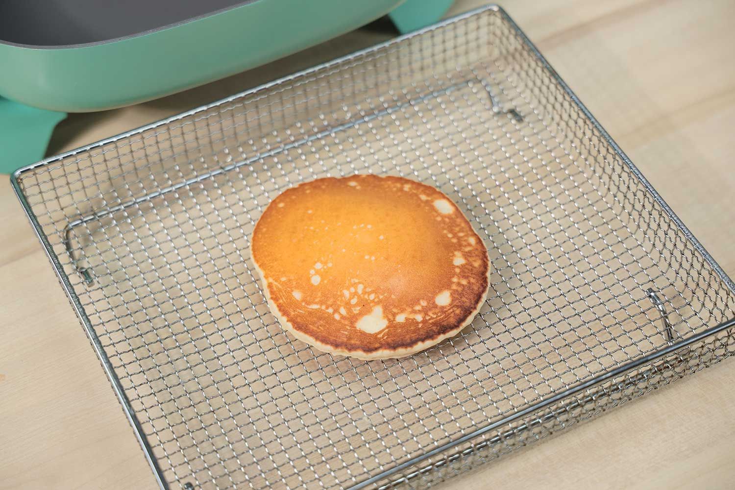 The top side of a golden brown pancake that’s dark around the edges inside an air fryer basket. In the corner is the GreenLife Ceramic Non-Stick Electric Skillet