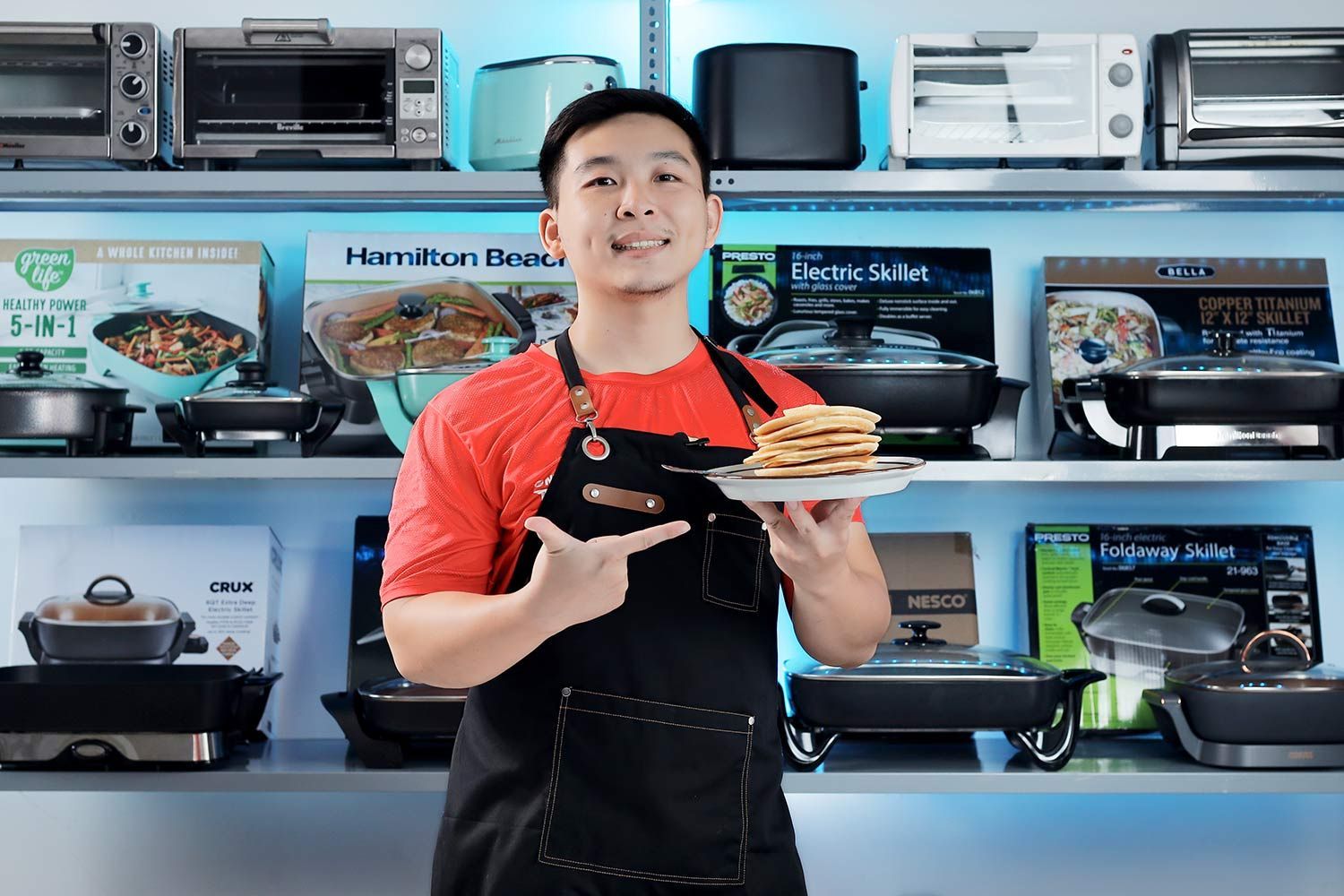 A person holding and pointing at a plate of pancakes. In the background is a shelf of electric skillets and toaster ovens.