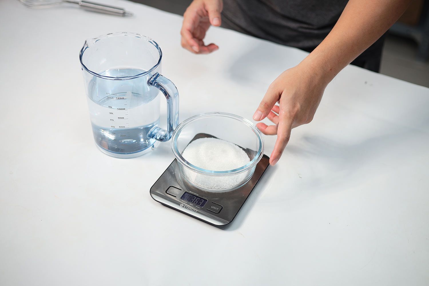 A measuring cup with 1 liter of water and a bowl of 363 g of sugar on a digital scale.