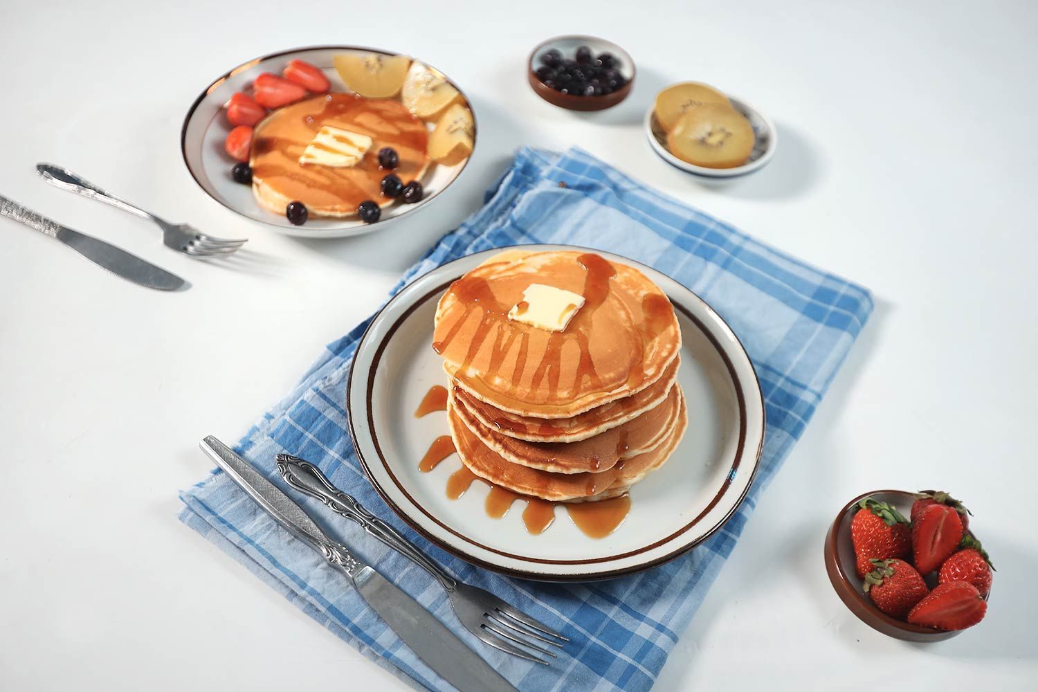 Two plates of pancakes with silver cutlery and small plates of blueberries, kiwis, and strawberries.