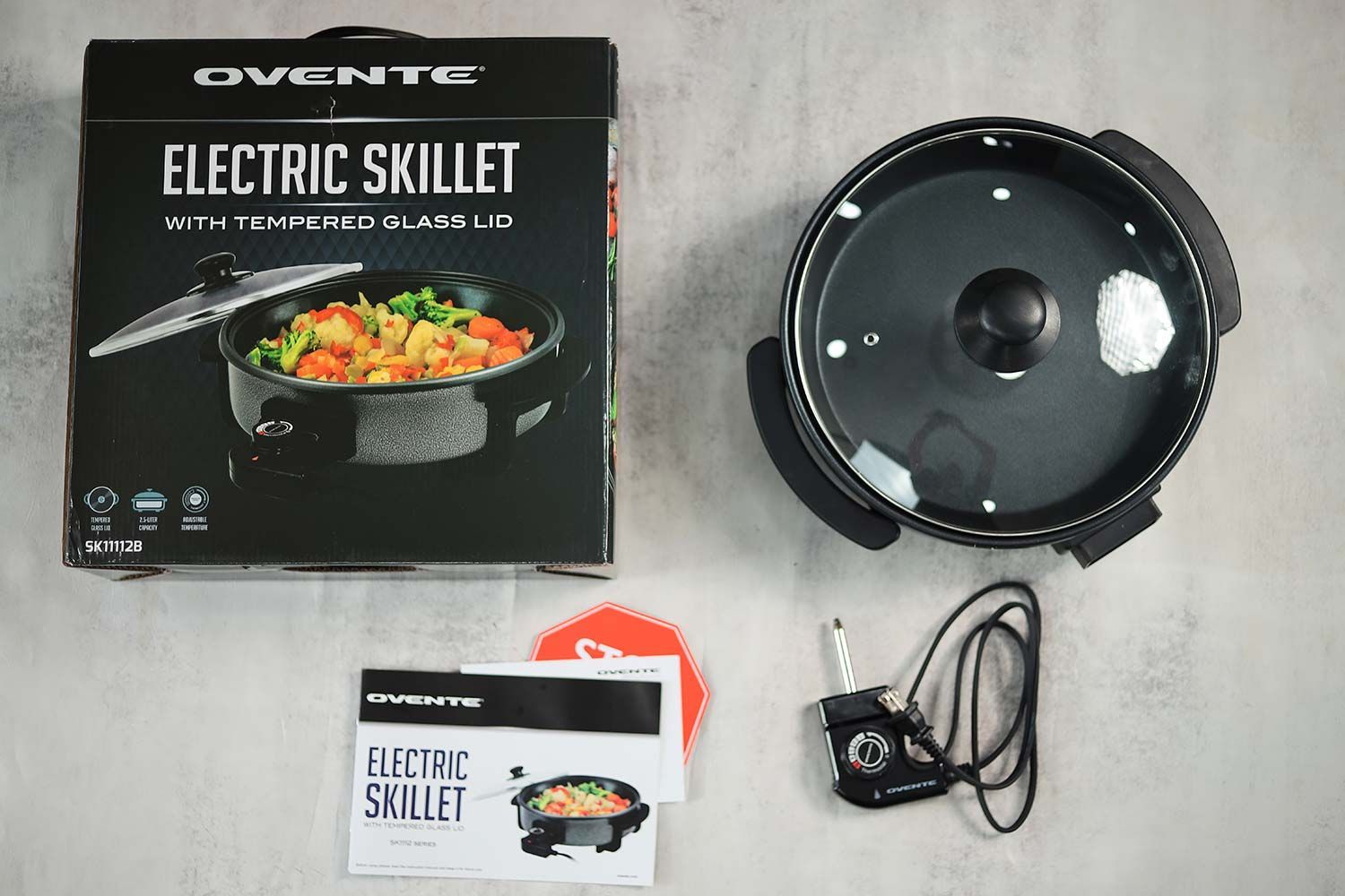 On the upper right is the Ovente Non-Stick Electric Skillet SK11112B. On the left is its cardboard box. Below the skillet, on the right is a thermostat and on the left is an instruction manual.